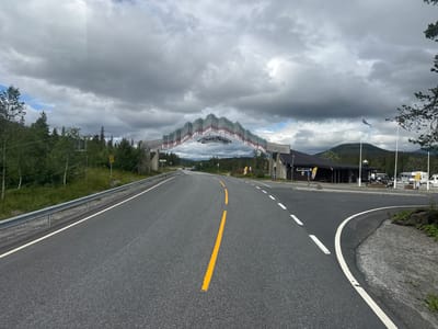 The image shows a road leading towards a large, decorative archway with the text "Nord-Norge" on it, suggesting it marks the entrance to Northern Norway. The archway is situated in a rural or semi-rural setting, with trees and hills visible in the background. There is a building complex to the right of the arch, which appears to be a roadside service area or a small village. Several flags are flying from poles near the building. The road itself is paved and has clear lane markings. The sky is overcast with dark clouds. The location is identified as Smalåsen, Norway. There are no identifiable people in the image.