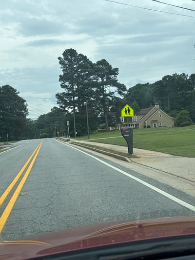 The image depicts a street in Mountain Park, Georgia, USA. The road is paved and marked with double yellow lines. A yellow pedestrian crossing sign is visible on the right side of the road, in front of a residential house. The sky is overcast. Trees line both sides of the road. Part of a car, possibly the vehicle from which the picture was taken, is seen on the right side of the frame.