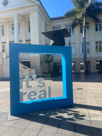 The image captures a sunlit outdoor scene in Kelvin Grove, Australia, with a classic building featuring columns and multiple windows in the background. In the foreground, a bright blue rectangular frame stands prominently, displaying white text on a transparent surface that reads "it's real". Behind the frame, a large mortarboard, a symbol of graduation, is positioned. A tall palm tree adds a tropical element to the environment. The ground is paved with square tiles, and the scene is characterized by strong shadows cast by the objects and the sun. No people are visible in the image.