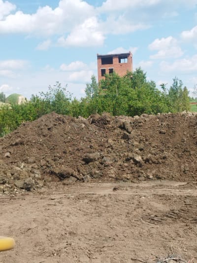 A brick building, likely under construction or in disrepair, stands amidst dense green foliage. The building, made of red bricks, has several window openings and appears to be missing a roof. In the foreground, a large pile of dark, earthy soil dominates the scene. The ground in front of the soil pile is bare, packed dirt with tire tracks visible, suggesting recent vehicle activity. The sky above is blue with scattered white clouds, indicating fair weather. The overall impression is one of an outdoor industrial or construction site, possibly in a rural or semi-urban area, with the natural elements of trees and soil juxtaposed against the man-made structure. The time of day appears to be daytime, given the bright lighting. The location is identified as Rozdil, Ukraine. No people or text are visible in the image.