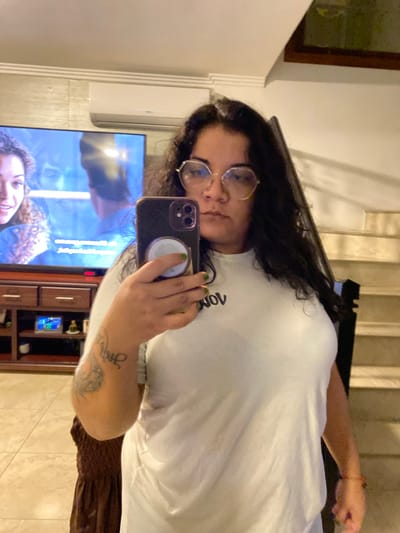 A person with dark curly hair and glasses holds a smartphone, taking a selfie. The individual is wearing a white t-shirt. Behind the person is a television playing content with visible figures. A dark wood cabinet sits below the television. The location appears to be indoors. The city of La Aurora, Ecuador, cannot be forensically determined from this image.