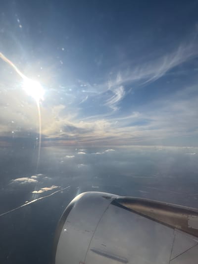 This media file presents an aerial view captured from the window of an airplane during daylight hours. The image is oriented sideways, but when viewed upright, it shows the sun on the left, the aircraft engine on the right, and the horizon stretching across the middle.
The sky dominates the upper and central parts of the frame, transitioning from a deep blue in the upper-left to lighter shades towards the horizon. Wispy, high-altitude clouds, characteristic of cirrus formations, are scattered throughout the blue expanse. Along the horizon, a denser band of clouds with hints of soft orange and pink suggests a low sun angle, possibly indicating early morning or late afternoon.
The sun is a prominent feature, located in the lower-left quadrant (when viewed upright). It shines brightly, causing significant lens flare and reflections on the aircraft window, including a distinct, multicolored streak of light extending horizontally from its glare. Numerous small, bright specks are also visible, likely dust or imperfections on the window surface.
On the right side, the robust structure of an aircraft's engine nacelle and a portion of its wing are clearly visible. The engine casing appears to be a light metallic grey or white, with visible rivets and panel lines. Part of the inner cowling or exhaust section reveals a metallic, golden-brown hue.
Below the horizon and the cloud band, a distant and indistinct landscape or body of water can be seen. It appears dark and hazy from the altitude, with faint, elongated light-colored streaks that are too ambiguous to identify as specific geographical features or structures.
There are no discernible people or readable text visible in the image. The scene conveys the experience of high-altitude travel on a day with varied cloud cover and bright sunshine. While the context of Combs-la-Ville, France, is provided, the image itself does not offer specific visual cues to confirm this geographical location.