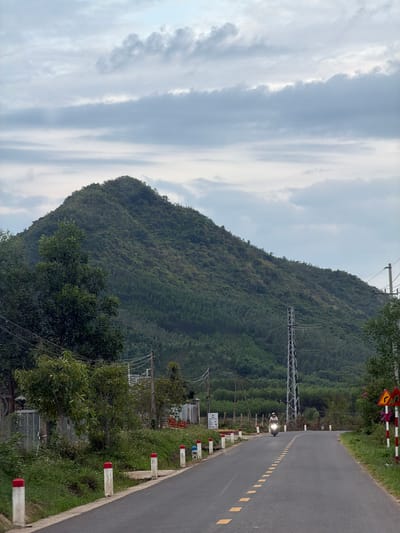 The image depicts a serene rural landscape on a cloudy day, likely in the city of Dien Tho, Vietnam. A paved road curves gently through the scene, marked by dashed yellow lines. A lone motorcyclist, wearing a helmet, travels away from the viewer. The roadside is lined with white bollards topped with red bands, and lush green vegetation. A prominent, tree-covered mountain dominates the background, its slopes densely wooded. A tall metal pylon stands to the right of the road, suggesting the presence of electrical infrastructure. Various trees, some appearing to be fruit-bearing, are visible along the left side of the road, interspersed with what looks like a fence and some simple structures. A traffic sign with a yellow background and a black arrow indicating a curve is visible on the right side of the road, next to more bollards. The sky is overcast with layers of gray and white clouds.