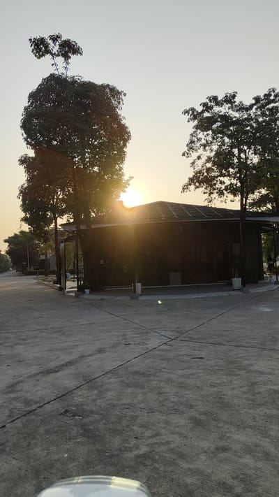 The image depicts a scene in the city of Kebalen, Indonesia, at what appears to be sunset or sunrise. The foreground is dominated by a concrete ground, possibly a road or a parking lot, with visible expansion joints forming a grid pattern. In the mid-ground, a dark-colored, modern-style building with a sloped roof is situated. The building features large windows, partially obscured by the sunlight. Two potted plants are placed near the building. Several trees are scattered around the scene. A prominent, large tree with dense foliage stands to the left of the building, with the sun peeking through its branches, casting lens flare. Another tree is visible on the right side of the building, also partially silhouetted against the bright sky. The background shows a street extending into the distance, lined with more trees and structures, suggesting a developed area. The sky is a pale orange and yellow, indicative of either dawn or dusk. The overall impression is of a tranquil, possibly residential or commercial, outdoor setting.