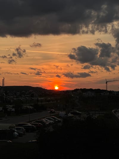The image shows a sunset over a residential area in Świebodzice, Poland. The sky is a vibrant orange and yellow, with dark clouds forming above. In the foreground, there is a street with parked cars and a few residential buildings. A construction crane is visible in the distance. There are no people visible in the image. The overall scene depicts a peaceful evening in a suburban environment.