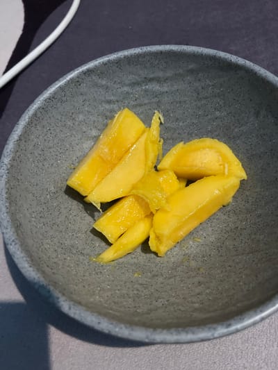 A close-up overhead shot shows several pieces of cut mango in a grey speckled bowl. The mango pieces are a vibrant yellow-orange color and appear to be fresh and juicy, with some glistening liquid visible. The bowl is slightly shadowed on the left side. The background consists of a dark grey textured surface with a portion of a white cord visible in the upper left corner. The image appears to be taken indoors, possibly on a table. No specific details about Bandarbireuen, Indonesia are discernible from the visual information in the image.