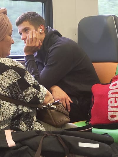 Two people are seated next to each other on a train. The person on the left, a woman, is wearing a headscarf and a black and white patterned top. Her left arm is resting on her lap. The person on the right, a man, is wearing a dark hooded sweatshirt and is looking towards the woman. He has a white earbud in his right ear and is resting his chin on his right hand. In front of the man is a red bag with white lettering that reads "arena". Various bags and objects are piled around the two individuals, suggesting a travel or commuting scenario. The background shows a train window with an out-of-focus view of greenery and a cloudy sky, consistent with a rural or suburban environment. The image was taken in the city of Rozhdestvenka, Russia.