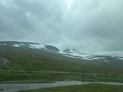 The image depicts a vast, natural landscape under an overcast sky. In the foreground, a shallow, rocky river flows across green grass. A thin, paved road runs horizontally across the middle ground, separating the river area from a sloping green mountainside. The mountain slopes are dotted with patches of snow, particularly towards the upper sections where rocky terrain and more extensive snowfields are visible. The sky is filled with thick, gray clouds, suggesting a cool, possibly damp, atmosphere. There are no people or man-made structures visible other than the road. The scene conveys a sense of remote, undeveloped wilderness.