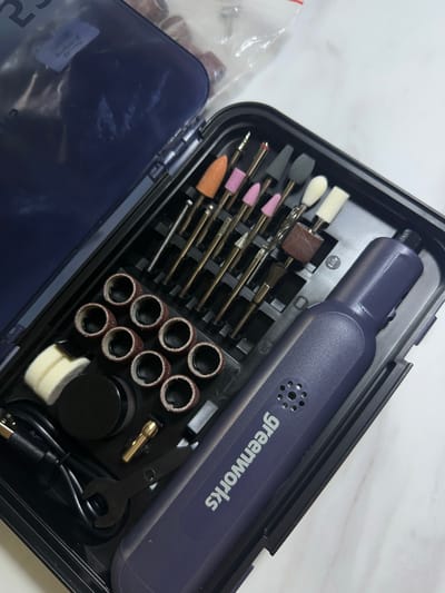 The image displays an open, dark-colored plastic storage case containing a compact rotary tool and numerous accessories. The main subject is a sleek, dark blue/purple handheld rotary tool, prominently featuring the "greenworks" logo in white text on its side. It has a textured grip and a perforated section near the logo, likely for ventilation.
The case's interior is meticulously organized with custom-fit compartments. To the left of the tool, there's an array of interchangeable bits, including various shapes and materials for grinding, sanding, and polishing. These include conical pink and orange bits, cylindrical brown bits, gray and white abrasive stones, and white felt-like polishing buffers. Several metallic engraving or cutting bits with fine points are also visible. Below these are cylindrical sanding bands, neatly stacked in rows.
Other accessories in the case include a small black wrench, a gold-colored collet, a coiled black USB charging cable, and a small black circular container. A clear plastic bag, partially visible on the far left, contains additional small items. The case's translucent dark blue lid is open and partially visible in the lower-left corner. The background appears to be a light-colored, possibly white or marble-patterned surface, suggesting an indoor setting. The items appear new and well-organized, indicating a complete kit ready for use. No specific location cues for Kuda, Russia, are discernible from the image.