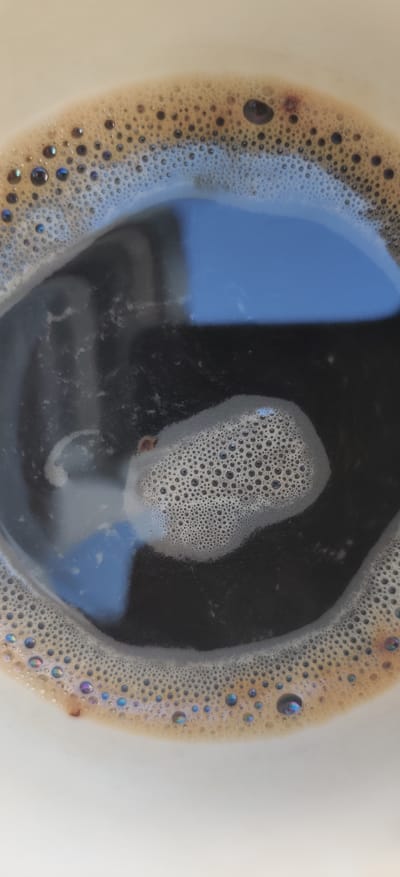 A close-up, overhead shot captures the foamy surface of a cup of dark coffee. The coffee has a rich brown color, and the foam is a lighter tan, creating a distinct border around the dark liquid. Small, iridescent bubbles dot the foam, reflecting hints of purple and blue. A larger, isolated patch of foam with denser bubbles sits in the center of the coffee. The coffee itself appears dark and reflective, showing a blurred blue and white reflection, possibly of a window or sky. The rim of the white ceramic cup is visible at the bottom and sides of the frame. The overall impression is one of a fresh, hot beverage. There are no people or identifiable landmarks in the image. The location in Rizunenkove, Ukraine, is not discernible from the visual information provided.