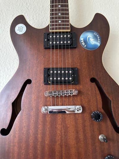 The image is a close-up, eye-level shot of the body of a brown electric guitar, likely a semi-hollow body model, against a textured white wall. The guitar is oriented vertically. The wood grain of the guitar is clearly visible. Two black humbucker pickups are mounted on the body. Chrome hardware, including a bridge and strap buttons, is also visible. On the upper left bout of the guitar, there is a circular sticker with text in German, reading "Gott gibt's echt!". On the upper right bout, there is a metallic blue circular sticker with a logo and the text "YWAM GERMANY". The guitar has a dark fretboard with white fret markers. A volume knob and a toggle switch are visible on the lower right side of the guitar's body. The scene is indoors and appears to be a still life arrangement, likely in a home or studio setting. There are no people present. There is no information in the image to indicate a specific location within the city of Bad Blankenburg, Germany.