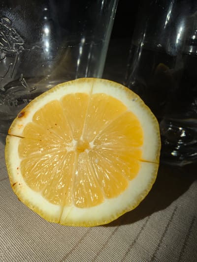 Kavrakirovo, Bulgaria: A close-up image presents a lemon slice, approximately half of a whole lemon, resting on a light-brown or beige textured surface with a discernible diagonal weave pattern. The lemon displays its yellow-orange pulp and white pith, with a minor cut or separation visible in the lower portion of the pith. Small, dark blemishes are present on the lemon's outer edge. In the blurred background, two clear glass vessels are visible. The glass on the left exhibits embossed decorative detailing, while the glass on the right is plainer. A shadow from the lemon is cast onto the surface towards the lower right. No individuals or active interactions are depicted.