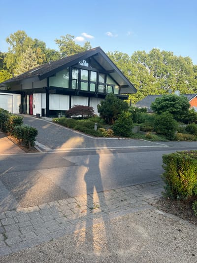 The image shows a modern house with a striking angular roof and extensive glass facade. The house is surrounded by lush green trees and well-maintained landscaping. A paved walkway runs alongside the house, separating it from a gravel path. The scene appears to be set in a residential area, possibly a suburb, given the presence of other houses in the background and the neatly manicured gardens.
The time of day is likely late afternoon or early evening, as indicated by the long shadows cast across the walkway and the warm, golden light bathing the scene. The sky is clear and blue, suggesting pleasant weather. The overall mood is peaceful and serene, with a sense of modern architectural design harmonizing with nature. There is no visible text in the image.