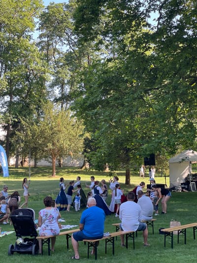 The image depicts an outdoor event taking place on a sunny day in Stupava, Slovakia. A group of people are gathered on a grassy area, with many seated on wooden benches or chairs. Some individuals appear to be participating in a structured activity, possibly a performance or class, as several are standing and engaged in synchronized movements. The setting includes trees providing shade, a stage area with speakers and equipment, and a tent in the background. The attire of the participants varies, with some in casual clothing and others in more formal or traditional outfits, suggesting a diverse audience or participants. A baby stroller is visible in the foreground, and a blue banner with text is present at the bottom of the image. The overall atmosphere appears relaxed and communal, with people enjoying the outdoor gathering. The time of day is likely afternoon, given the sunlight and shadows.
