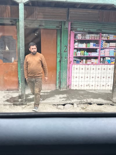 A man walks in front of a shop in Dras, India. The man is wearing a brown sweater and brown pants. He has a beard and is looking to the side. The shop is painted pink and green and has shelves stocked with various products. The ground in front of the shop is wet and muddy. The scene appears to be a street-level view, possibly from inside a vehicle, looking out.