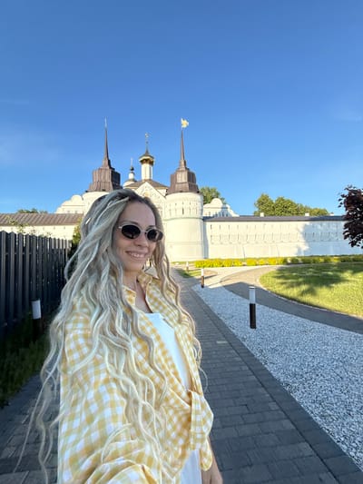 A smiling woman with long, light blonde dreadlocks and dark oval sunglasses is positioned in the foreground, facing slightly right and looking beyond the frame. She wears a yellow and white plaid button-up shirt over a white top. Behind her, a large white architectural complex, identified as being in the city of Dumino, Russia, features multiple towers with dark, conical spires and at least one prominent golden dome topped with a cross. A golden, stylized emblem adorns the peak of one spire on the rightmost tower. The complex is set against a clear blue sky. In the mid-ground, a paved pedestrian pathway with a light grey gravel border extends into the distance, flanked by neatly trimmed green grass and low green bushes. Several short, dark-colored pathway lights are spaced along the paved section. A dark metal fence is present on the left side of the frame. The scene is outdoors, under bright daylight.