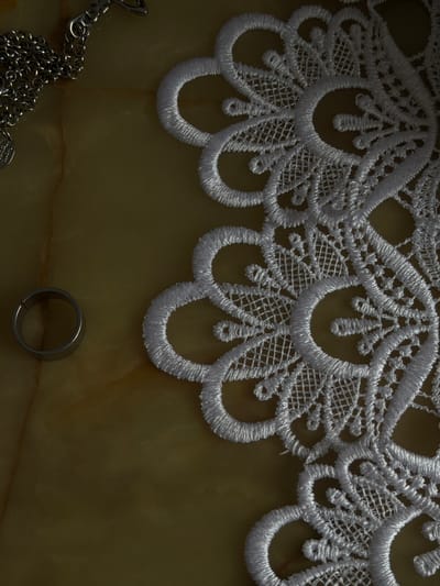 A close-up, top-down shot shows a segment of white lace bordering the top of a translucent, yellowish-green surface with marbled patterns. The lace features a scalloped edge with intricate, geometric and floral designs created with white thread. Below the lace, a silver-colored ring with a simple, band design is positioned in the lower right quadrant of the image. To the left of the ring and extending towards the bottom left corner, a portion of a silver chain is visible, with some of its links and attachments creating a tangled pattern. The lighting appears to be from the side, casting shadows that highlight the textures of the lace, the marbled surface, and the metallic sheen of the chain and ring. The setting is likely indoors, with the surface possibly being a stone countertop or a decorative table. No people, text, or specific time of day or weather cues are discernible.