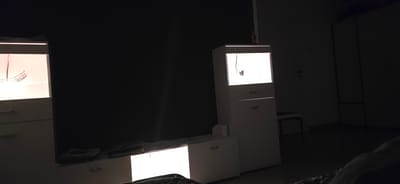 In a dimly lit room, likely in Villing, France, two white illuminated cabinets are visible. The cabinet on the left has a lit interior displaying a curved metallic object. The cabinet on the right is taller, with a lit top compartment showing a small object, and two drawers below. A long, thin, dark object hangs from the ceiling towards the right cabinet. In the background, a part of a wardrobe or closet is visible, along with a stool. The floor appears to be dark. The overall impression is one of a domestic interior, possibly a living room or bedroom, with an emphasis on the illuminated display cabinets.