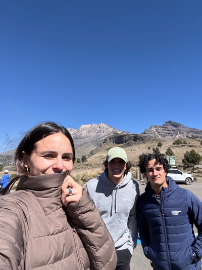The image depicts three individuals posing for a photograph in a mountainous, high-altitude outdoor environment identified as Tlamacas, Mexico, under a clear blue sky.

In the foreground, a female with dark hair is positioned on the left, smiling directly at the camera. She wears a dark brown puffy winter jacket, the zippered collar of which she holds up with her right hand, revealing a silver ring on her ring finger and a silver hoop earring on her visible left ear. To her right stands a male with dark curly hair, smiling. He wears a light green baseball cap with a white "NY" logo and a grey hooded sweatshirt with a white Nike logo. To his right, another male with dark curly hair looks towards the camera with a neutral expression. He wears a dark blue puffy winter jacket with a white "SCANDINAVIAN EXPLORER 72°38'21" logo on the left chest. Both male subjects have their hands in their pockets.

The background features rugged mountain terrain. A prominent snow-dusted or light grey rocky mountain peak dominates the central background, flanked by lower, rocky, and treeless peaks to its right. The mid-ground consists of dry, golden-brown grass and scattered small coniferous trees. A paved or dirt road section is visible on the far right, where a white SUV or crossover vehicle is parked. Behind the vehicle, a light-colored structure, possibly a sign or small building, is discernible. A person in a blue jacket is partially visible on the far left, walking away from the camera. The sky is uniformly clear and bright blue. Three small, faint magenta circular artifacts are present in the sky area.