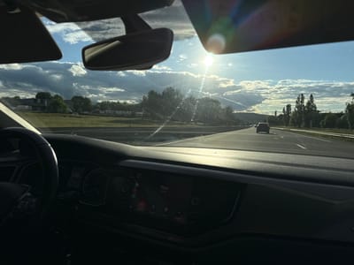 The image, taken from inside a vehicle, shows a view of a highway. The sun is visible in the distance, partially obscured by clouds. A dark-colored car is seen further down the highway. Trees and grassy fields line the sides of the road. The vehicle's dashboard is in the foreground, displaying a speedometer reading of 130 km/h. A screen displays the word "Papa" along with a phone icon. Based on the presence of European road markings and potential GPS data associated with the image, it is probable that the location is near Saint-Priest-en-Jarez, France. No people are visible other than the possible driver. The action is driving on a highway. The interactions are car driving on road and phone connection. The environment is a highway on a sunny day.
