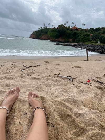 This is a photograph taken from a low angle, looking down at a person's feet resting on a sandy beach. The image is oriented sideways, with the ocean and a cliffside to the left and the sand and feet occupying the majority of the frame to the right.

The subjects are a pair of bare feet with toenails painted white and adorned with shell anklets. One foot has a silver ring on the second toe. The sand is a light beige color and appears slightly damp in places, with small pieces of driftwood, shells, and debris scattered across it. A small red object, possibly a piece of plastic, is visible in the sand near the center of the image.

The scene is a beach in Batawala, Sri Lanka. To the left of the frame, the ocean is visible with waves breaking on the shore. The water is a pale turquoise color. Beyond the beach, a lush green cliffside rises, dotted with palm trees and red-roofed buildings. The sky is overcast and gray, suggesting cloudy weather, possibly approaching rain.

The overall mood of the photograph is relaxed and peaceful, with the focus on the simple pleasure of being at the beach. The time of day is unclear due to the overcast sky, but the lighting suggests it is daytime. There is no visible text in the image.