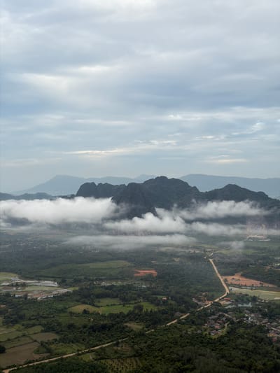 The image is an aerial landscape view from an elevated vantage point overlooking a rural area. In the foreground, dense green foliage covers rolling hills and terraced agricultural fields. A dirt road winds through the landscape, leading towards a cluster of buildings that form a small village. Beyond the village, more agricultural land, including rice paddies, is visible. In the middle ground, a range of dark, tree-covered mountains rises, partially obscured by a thick layer of white clouds or mist that snakes between the peaks. Further in the distance, additional mountain ranges are visible, fading into a hazy, muted blue against a cloudy, overcast sky. The overall impression is one of a serene, undeveloped rural environment, possibly Hong Ngern Village, Laos, characterized by its mountainous terrain, lush vegetation, and agricultural activities.