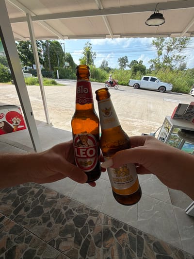 In Bang Muang, Thailand, two hands hold up beer bottles in a first-person perspective. One hand, with visible knuckles and forearm hair, holds a dark glass bottle labeled "LEO LAGER BEER" with a leopard logo. The other hand, with manicured fingernails, holds a lighter glass bottle labeled "SINGHA THE ORIGINAL THAI BEER ESTD 1933." The bottles are angled towards each other. The background shows a paved area with stone-like tiling, leading to a road. A white pickup truck is parked on the road, and a person on a motorbike is riding past. Lush green vegetation borders the road, and trees are visible under a bright blue sky with scattered clouds. A white awning provides shade over the tiled area where the photo is taken. A discarded container with images of ice cream cones is visible to the left.