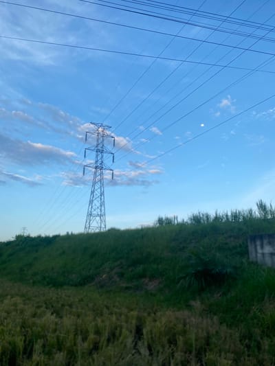An image of an electrical transmission tower. The tower is constructed of a steel lattice structure and stands beside a grass-covered slope or embankment. Electrical lines are strung from the tower and reach to other unidentified points. In the background, there's a sky that is partly blue with some scattered clouds. There may be a visible moon in the sky. The image orientation is sideways. The location is identified as Payarangkuluh, Indonesia, though no landmarks are visible to specifically confirm the place. The scene lacks any indication of human presence or activity.