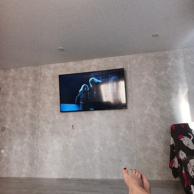 The image shows a modern living room with a large television mounted on a textured gray wall. The television displays a scene from a movie or TV show, featuring two individuals in a dimly lit setting. Text is visible on the screen, partially obscured by reflections, but "ON THE SPOT" and "WORK PROGRAM" can be discerned.
In the foreground, a person's bare foot with red nail polish is visible, suggesting a relaxed and casual atmosphere. The room appears to be well-lit by natural light coming from the right side of the frame, casting a bright glow on the wall and part of a dark-colored armchair with a pattern of white cats and red hearts. The floor is covered with light gray laminate flooring.
The overall impression is one of a cozy evening at home, with someone enjoying entertainment on television. The scene is indoors, likely in a residential setting in Podlubovo, Russia. No specific time of day or weather conditions are evident. The mood is peaceful and domestic.