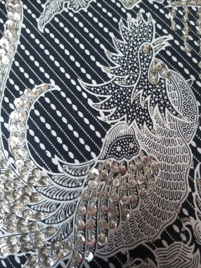A close-up shot reveals a fabric with an intricate batik pattern in black and white. The pattern depicts a stylized bird or phoenix motif with detailed feather designs and surrounding decorative elements. Silver sequins are meticulously attached, highlighting the outlines of the design and adding a sparkling, textured effect. The background features diagonal lines of small white dots against a dark background.