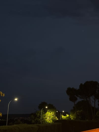 The image depicts a nighttime scene with a dark, overcast sky. In the foreground, a neatly trimmed hedge runs horizontally across the lower portion of the frame. Behind the hedge, several streetlights illuminate the surrounding trees and foliage. On the left, a tall, white lamppost with a single light shines brightly. To the right, another, shorter lamppost is nestled amongst dense, brightly lit green trees. Further in the background, silhouetted trees and more distant streetlights suggest a populated area. A faint, distant town or village is visible on the horizon. In the upper right corner of the sky, a tiny celestial body, possibly a star or planet, is faintly visible. The overall impression is of a tranquil evening on the outskirts of El Escorial, Spain.