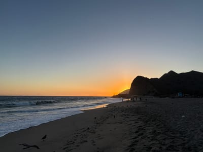 At sunset, the sky over the city of Old Malibu, United States of America, transitions from a light blue at the top to a vibrant orange glow near the horizon. The sun is partially obscured by a dark, silhouetted mountain range, creating a sunburst effect. A beach with dark sand stretches across the foreground, with footprints visible. Waves crash onto the shore, with white foam receding on the wet sand. Several seagulls are present on the beach. In the middle ground, a group of people are silhouetted against the bright sky as they walk along the shoreline. A small, light blue lifeguard tower is visible in the distance, near the base of the mountains.