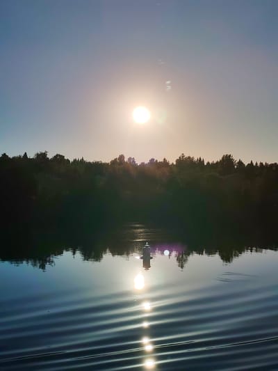 A natural landscape scene captured in Ivanov Bor, Russia, at what appears to be a low sun angle, likely sunrise or sunset. The upper third of the image is dominated by a clear to lightly hazy sky, transitioning from a warm, pale yellow-orange tone near the sun on the right to a cooler desaturated blue-grey towards the upper left. A bright, white, circular light source, presumably the sun, is prominently positioned in the upper-right quadrant of the sky, emitting visible light flares and a strong luminosity.
Below the sky, a dense, dark forest line stretches across the horizon, appearing as a silhouette against the brighter sky, with various tree types discernible. The lower two-thirds of the image consist of a calm body of water. The surface of the water displays a strong, vertical reflection of the sun, rendered as a series of distinct, elongated, bright white ovals due to surface ripples. Located in the lower-middle section of the water, slightly to the left of the sun's reflection, is a solitary white cylindrical object, appearing to be a buoy or navigational marker, which also casts a direct reflection beneath it. The water in the foreground, particularly towards the lower left, exhibits clear, parallel ripples. Reflections of the dark forest are visible as a distorted band beneath the actual tree line on the water's surface. No individuals or overt human activities are present in the image.