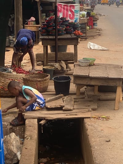 The scene depicts an active, informal street market in AlakIa, Nigeria. In the immediate foreground, an adult female, barefoot, dressed in a blue patterned top and headwrap, is bent over, sorting a large pile of red chili peppers contained within a large woven basket on the ground. Adjacent to her, a young boy, wearing a multi-colored sleeveless shirt and dark shorts with "GUCCI" branded slides, sits on a wooden plank covering part of an open concrete drainage ditch. He is leaning forward, manipulating a piece of wood or debris over the dark, refuse-filled water in the ditch.
Behind the female, a raised wooden stall displays multiple small green bowls stacked with red tomatoes or peppers. A white banner with red "POS" text is prominently displayed above this stall. Below the stall, on the dirt ground, are a black plastic bucket, a brown bucket, and several rocks. To the right, another rustic wooden table sits beside the ditch, holding a gray speckled bowl and a green bowl. The ground consists of uneven dirt and gravel, with scattered wooden planks and debris. In the background, a paved road is visible, with blurred figures of pedestrians, multiple motorcycles, and a yellow auto-rickshaw indicating ongoing street traffic. Additional market stalls with produce, including watermelons, are visible further down the street.
