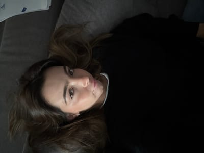 A single female individual is depicted lying supine on a dark gray upholstered surface. Her head is positioned towards the upper left of the frame, with her body extending diagonally downwards. She possesses long, wavy brown hair spread around her head and light brown eyes, which are directed towards the camera. The subject is wearing a black long-sleeved top, with a white collar visible at the neck. A white, folded object is discernible in the upper left background. The scene is an indoor environment in Nogueira e Silva Escura, Portugal.