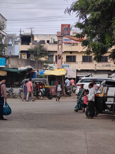 The image depicts a street scene in Malkapur, India, likely near a market or commercial area, with buildings lining the background and vehicles and pedestrians occupying the foreground. In the center of the frame, a yellow and black auto-rickshaw is parked, with a man wearing a red t-shirt driving it. Several people are visible around the auto-rickshaw and on the street. To the left, a man in a brown shirt and dark pants carries a blue duffel bag. Two women, one in a blue saree and the other in a pink outfit, walk past a parked motorcycle. Further to the right, a white SUV is parked, and a group of people, including a woman in a green saree and a child, stand near it, along with a man in a white shirt and dark pants. The street is paved with asphalt, and there are several shops and stalls with awnings. Overhead power lines crisscross the sky, which is overcast. A large tree with green foliage is present on the right side of the image, partially obscuring the buildings. The architecture of the buildings in the background appears to be a mix of traditional and more modern styles. A billboard with an image of a deity and text in an Indian script is visible on one of the buildings.