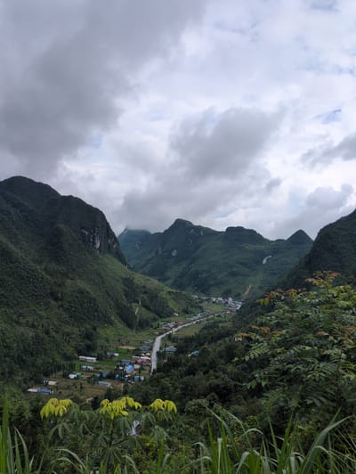 The image shows a wide, green valley in Phố Bảng Commune, Vietnam, under a cloudy sky. The valley is surrounded by lush, tree-covered mountains on both sides. A road snakes through the valley, with a small village situated alongside it. The village consists of numerous houses with colorful roofs, interspersed with patches of cultivated land. In the foreground, tall grasses and leafy branches frame the view, adding depth to the scene. The overall impression is one of a tranquil, rural landscape.