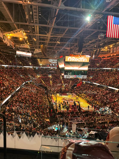 The image shows a packed basketball arena in Cleveland, United States of America. The American flag hangs from the ceiling amidst metal beams and lights. Large video screens display advertisements, including "Rocket Mortgage FieldHouse" and "Redfin". The basketball court is crowded with people, likely players and staff. The stands are filled with spectators.