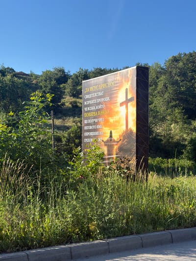 A large, rectangular billboard featuring a religious image is prominently displayed outdoors. The billboard is positioned at an angle, with its right side closer to the viewer. The image on the billboard depicts a sunset or sunrise over a hill, with a large cross silhouetted against the bright sky. A figure of Jesus Christ, arms outstretched, is visible at the base of the hill. Text is present on the billboard in what appears to be Cyrillic script.
The billboard is surrounded by lush green vegetation, including tall grasses and bushes. The foreground shows a grey concrete curb and pavement, suggesting a roadside location. In the background, there are trees and a hillside, characteristic of a rural or semi-rural environment. The sky is clear and blue, indicating fair weather.
The scene is located in the city of Veselinovo, Bulgaria, as indicated by the context provided. The overall impression is one of a roadside religious advertisement set against a natural landscape.
