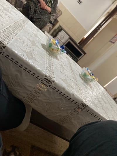 This image depicts an indoor domestic scene in the city of Branichevo, Bulgaria. A rectangular table, covered with a white, textured tablecloth featuring embossed patterns, dominates the foreground. Two clear glass bowls, each containing various brightly wrapped candies, are positioned on the tablecloth.
In the upper left background, a seated individual is partially visible from the mid-torso down. This person is wearing dark, patterned clothing, and their hands are clasped together in their lap. Directly behind the table in the upper right background, a dark brown furniture item, resembling a cabinet or stove, is situated against a light-colored wall, which exhibits visible tiling in its upper section. A wall-mounted fixture is present high on the wall in the distant background.
The lower foreground of the image shows the legs of an individual, clad in dark trousers. A white shoe is visible on the left foot, indicating the perspective of the photographer or another person seated at the table. The floor beneath the table is covered with a light-colored, possibly beige, carpet or rug. A dark brown table leg is visible beneath the tablecloth in the lower-center.