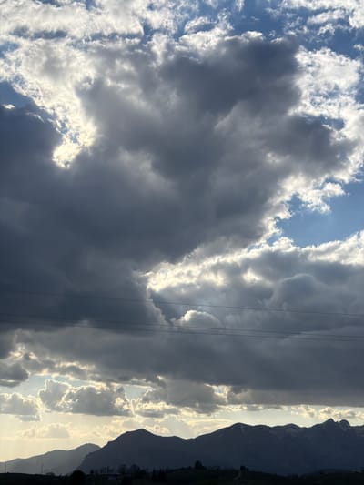 The image shows a sky filled with dark, heavy clouds interspersed with patches of sunlight. Mountains are visible on the right side of the frame, silhouetted against a brighter horizon. Thin power lines cut across the sky. The scene is of the city of Pišče, Montenegro.