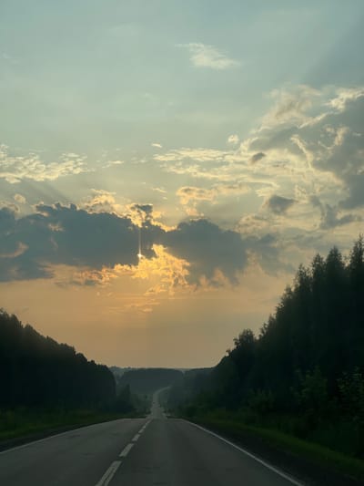 The image depicts a scenic view of a road leading through a forest in Novaya Kazmaska, Russia. The sun is setting, casting a warm glow over the landscape. The sky is filled with dramatic clouds, with rays of sunlight peeking through. The road is paved and appears to be in good condition, with a white line marking the center. Dense green trees line both sides of the road, creating a tunnel-like effect. In the distance, the road curves and disappears into a hazy horizon, suggesting a long and winding journey ahead. The overall impression is one of tranquility and natural beauty.