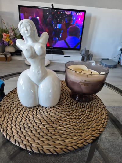 On a round glass table sits a woven placemat, a "Gingered Amber" candle in a glass jar, and a white ceramic sculpture of a woman's torso. In the background, a television displays a colorful image. Flowers are visible beneath the sculpture. The image was taken indoors, possibly in a living room setting. No information suggests that the image was taken in Villerupt, France.