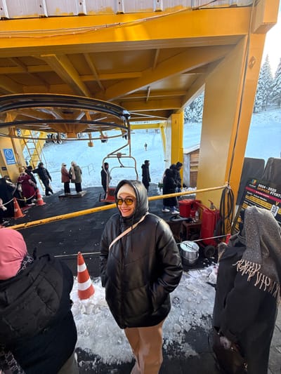 The image depicts a winter scene at the Uludağ ski resort in Bursa, Turkey, specifically at a ski lift station. The dominant feature is a large, yellow metal structure of a ski lift terminal, complete with a prominent pulley wheel and several attached chairlift seats. The ground beneath the station is a dark, partially snow and ice-covered surface. Snow-covered evergreen trees are visible in the background, along with a snowy slope where several small figures appear to be skiing or snowboarding.

In the foreground, a woman stands centrally, smiling towards the camera. She wears a black hooded puffer jacket, light-colored beanie, yellow-tinted sunglasses, and light-colored trousers, with a light-colored strap diagonally across her chest. Her hands are in her pockets. To her immediate left, a person is partially visible from the back, wearing a pink head covering and a dark quilted jacket. To her right, another person is partially visible from the back, wearing a dark coat and a grey patterned scarf with fringe.

In the mid-ground, under the yellow structure, multiple individuals in winter attire are gathered on a platform, appearing to be queuing for the ski lift. Two orange traffic cones are positioned on the snowy ground, partially embedded in ice. To the far right, a dark sign with white and green text displays "ULUDAG 2024-2025 SKI PASS FİYATLARI." Adjacent to this sign, near a small wooden shack, red fire extinguishers, hoses, and other utility items are visible on a small wooden stand or cart. A silver metal container rests on the snowy ground nearby. A blue circular sign with a white symbol is affixed to a yellow post on the left side of the station.