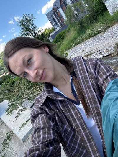 A young woman with brown hair and blue eyes is captured in a selfie, tilting her head slightly to the left as she smiles gently. She is wearing a brown and white plaid shirt over a white v-neck top and a silver necklace with a dark pendant. Her hair is pulled back into a braid. The background suggests an outdoor setting near a river or stream, with stone embankment lining the water. Green foliage and trees are visible, as well as a modern building in the distance. The lighting indicates it is daytime, possibly late afternoon or early evening, with scattered clouds in a blue sky. The image appears to be taken in the city of Kolašin, Montenegro, given the context provided.