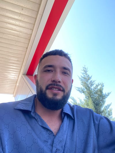 A man with a beard smiles at the camera. He is wearing a blue button-up shirt with a repeating pattern. Behind him, a tree is visible against a clear blue sky. The scene appears to be outdoors, under a roofed structure with a red and white striped awning. The overall impression is a candid snapshot taken on a sunny day.