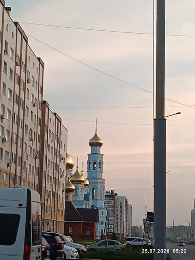 The image depicts a street scene in Mariinsky, Russia, at dawn, as indicated by the timestamp "25.07.2026 05:22". In the foreground on the left, a white van is parked. To its right, several cars are parked along the street. In the middle ground, a light blue and white church with golden domes is prominently visible. Adjacent to the church, a long, multi-story residential building with many windows runs along the left side of the street. Further down the street, more residential buildings and trees line the road, with a street light on a tall pole dominating the right side of the frame. The sky is overcast with soft, pastel colors, suggesting the early morning light. The overall atmosphere is calm and quiet.