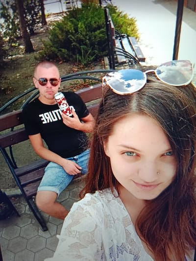 A man and a woman are seated on a park bench. The man, bald and wearing sunglasses and a black t-shirt, holds a can of soda. The woman, with long brown hair, is in the foreground, her face partially visible. They are in an outdoor setting with trees and bushes behind them. The ground is paved with hexagonal tiles. The scene appears to be in Pishchane, Ukraine.