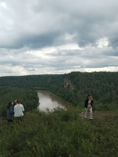 This image depicts a scenic overlook in Idrisovo, Russia, featuring a winding river bordered by dense, green forest. The sky is overcast with dramatic clouds. On the left side of the frame, three individuals are gathered on a grassy knoll, their backs mostly to the camera. One person is wearing a dark jacket, another a light-colored jacket, and the third's attire is less distinct. To the right, a woman stands on a dirt path, wearing a white crop top and light-colored flared pants, with a dark jacket draped over her shoulders. She is looking away from the camera and appears to be holding a phone. The foreground is filled with tall, green grass and some small white and yellow flowers, suggesting a natural, undeveloped area. The overall scene conveys a sense of exploration and appreciation for the natural landscape.