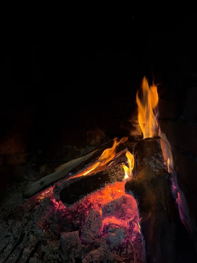 A dimly lit image showing burning wood in a fireplace. The fire is concentrated on the right side of the frame, with flames and glowing embers. Some flames appear to be swirling and elongated. The left side of the image is mostly black, suggesting a shadowed interior. The composition emphasizes the contrast between the bright flames and the dark surroundings. The location in Lilkovo, Bulgaria is not discernible from the image.